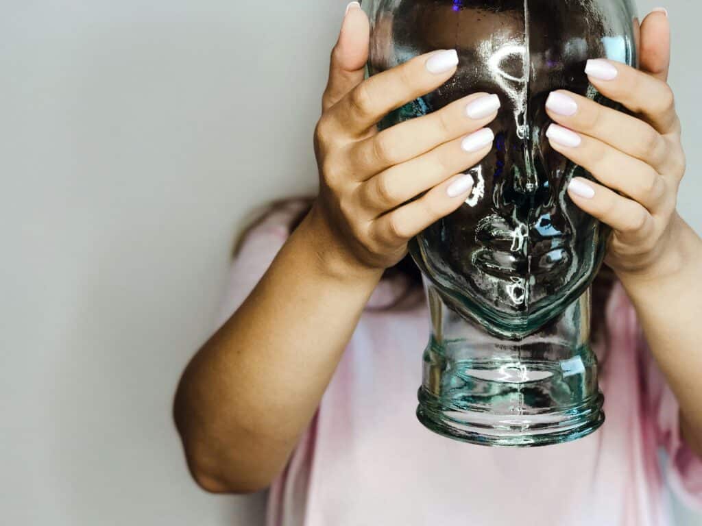 A woman hides behind a glass head figurine, using her hands as a shield indoors.